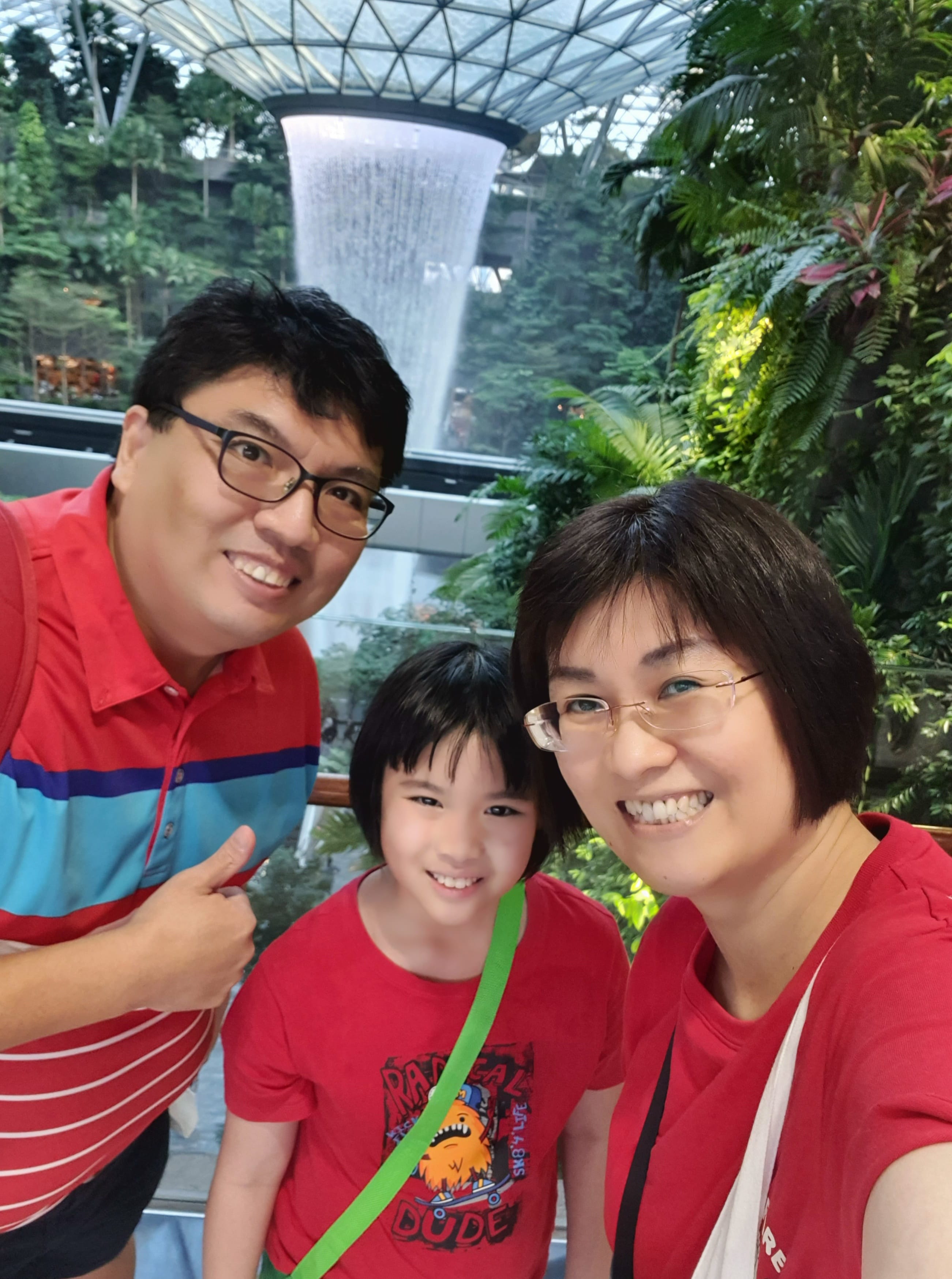 Three smiling people in red shirts pose indoors near a large indoor waterfall surrounded by lush green plants.